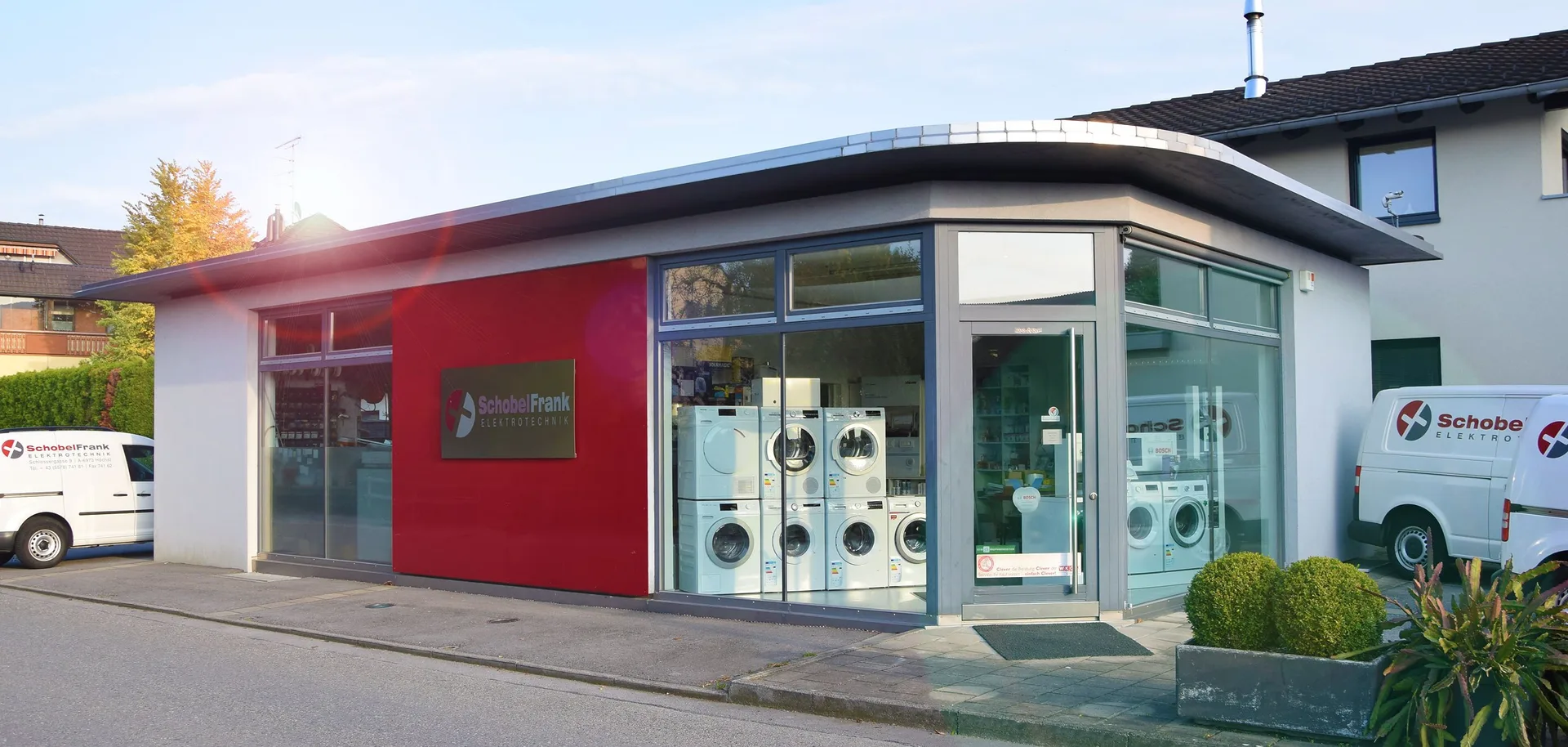 Das Betriebsgebäude von Schobel Frank Elektrotechnik in der Abendsonne, rote Fassade mit beleuchtetem Firmenschild, Schaufenster mit Hausgeräten, links und rechts ein beschrifteter Firmenwagen