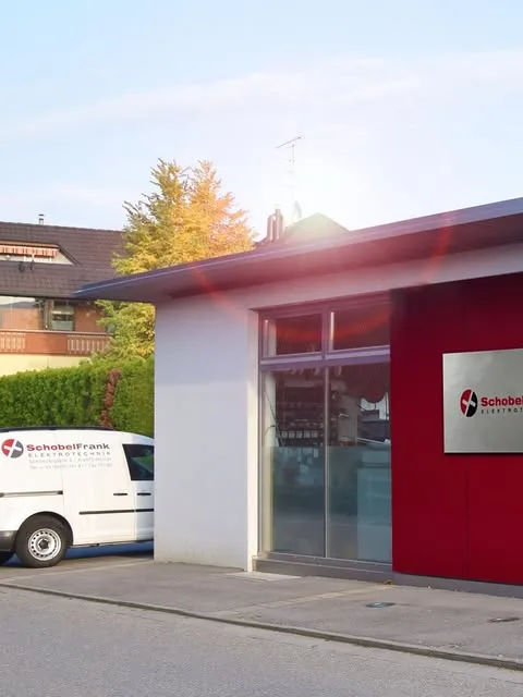 Die Gebäudeecke mit dem Firmenschild an der roten Wand, davor der Firmenwagen mit Logo und Adresse