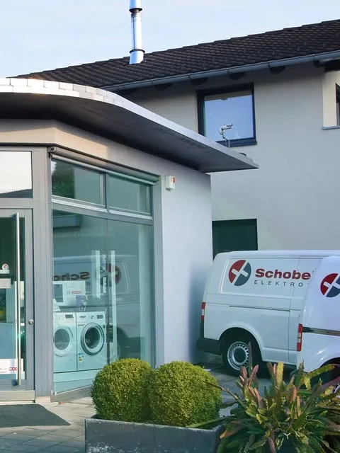 Eingangstür und Schauraum von der Seite, davor der Firmenwagen mit dem Logo von Schobel Frank Elektrotechnik